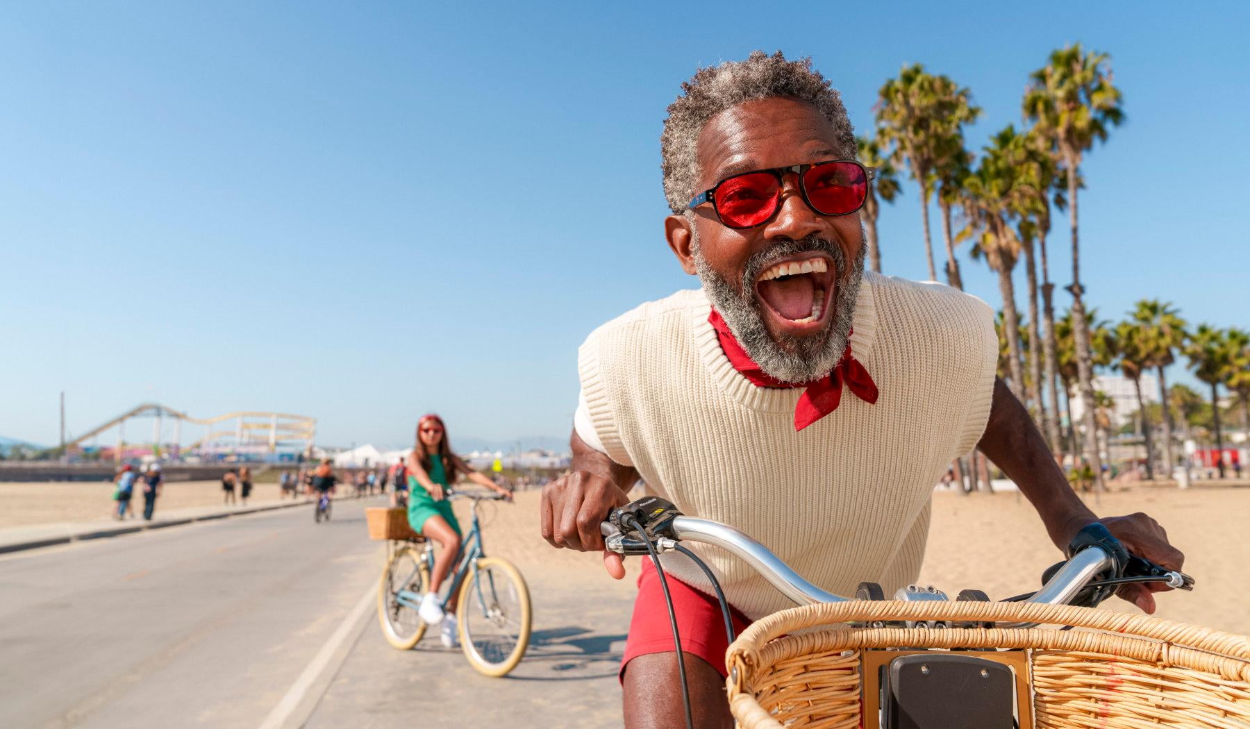 A photo of a happy man on a bicycle, wearing red sunglasses, outdoors with palm trees in the background.